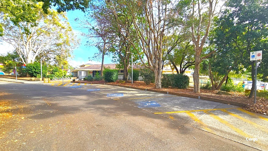 Accessible parking spaces at Cleveland Aquatic Centre are marked by a blue accessible parking sign separated by diagonal yellow lines and between diagonal yellow lines at the end. It also has a mobility disability logo parking sign, and a no-stopping sign. Tall trees with abundant foliage are lined within the garden full of dried leaf litter.