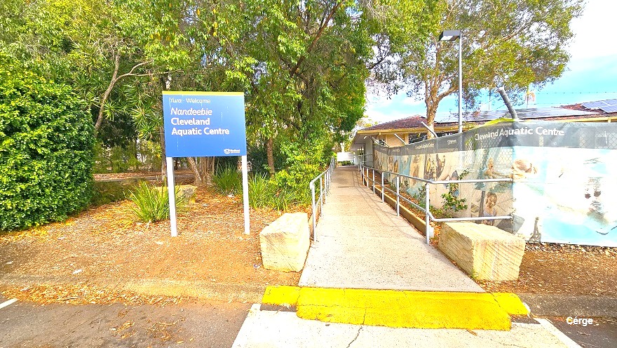 Smooth pathway with steel handrails leading to the entrance of Cleveland Aquatic Centre. At the start of the pathway is a wide yellow paint and two large rectangular rocks. On the right is a medium size blue signboard detailing the name of the center with bushes, shrubs, and trees behind. On the right is a fence fully covered by a large advert poster.