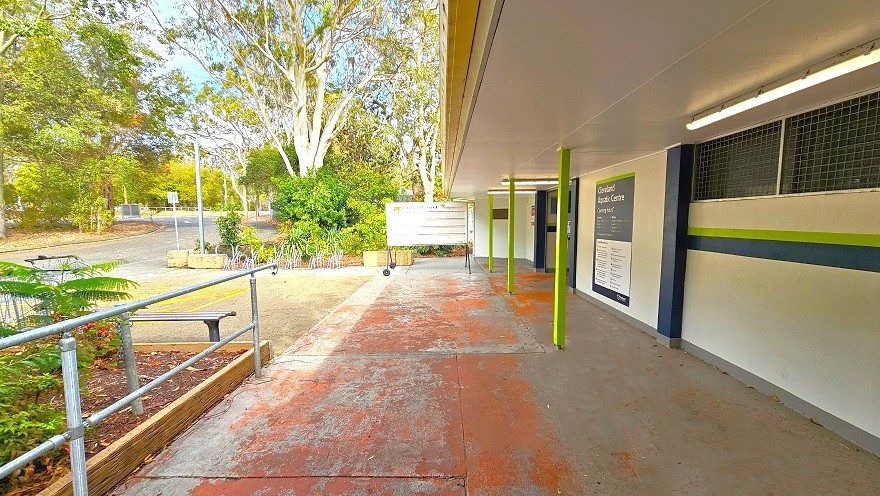 Free step access just before the entrance hallway of Cleveland Aquatic Centre has red floor markings. Before the entrance is a mobile whiteboard with black wheels. The front wall of the facility is white-painted with blue and lime-green features with a small metal grille. The ceiling is white with lime green narrow foundations. The bike rack and road is visible from this position.