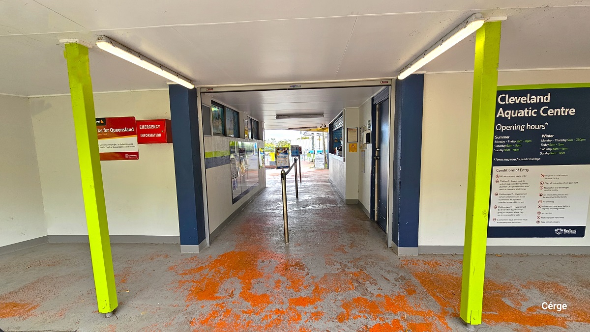 Entryway between two buildings with a low white ceiling. It has red floor markings and middle rope bollards to regulate traffic inside and outside of the centre. The entryway has lime green narrow foundations. The opening hours of the centre is detailed on the right white wall within a large blue, white, and lime green signboard. On the left wall is a red emergency information box.