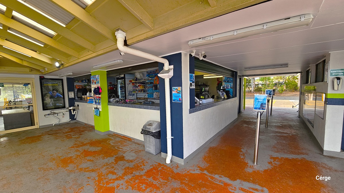 Reception, retail, and a small cafe at Cleveland Aquatic Centre. These amenities are within the left building with a white wall and blue and lime green foundation. In front of the retail counter is a grey garbage bin with a black garbage bag. The ceiling has yellow roof beams. On the right wall is a sanitization station above the blue and lime green paint.