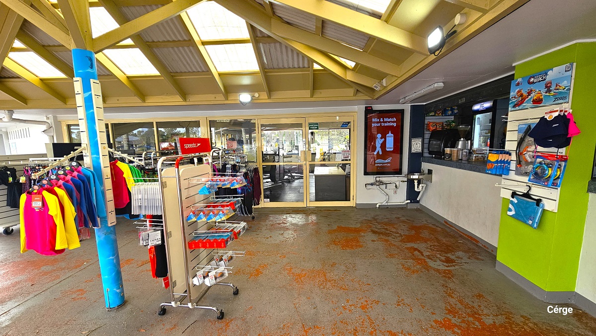 A closer look at the retail section next to the reception area. It has swimwear, goggles, and other swimming accessories. The ceiling has yellow beams with a skylit roof cover. The floor has red markings. After the retail and small cafe is a manually operated glass door with cream-colored glass frames.
