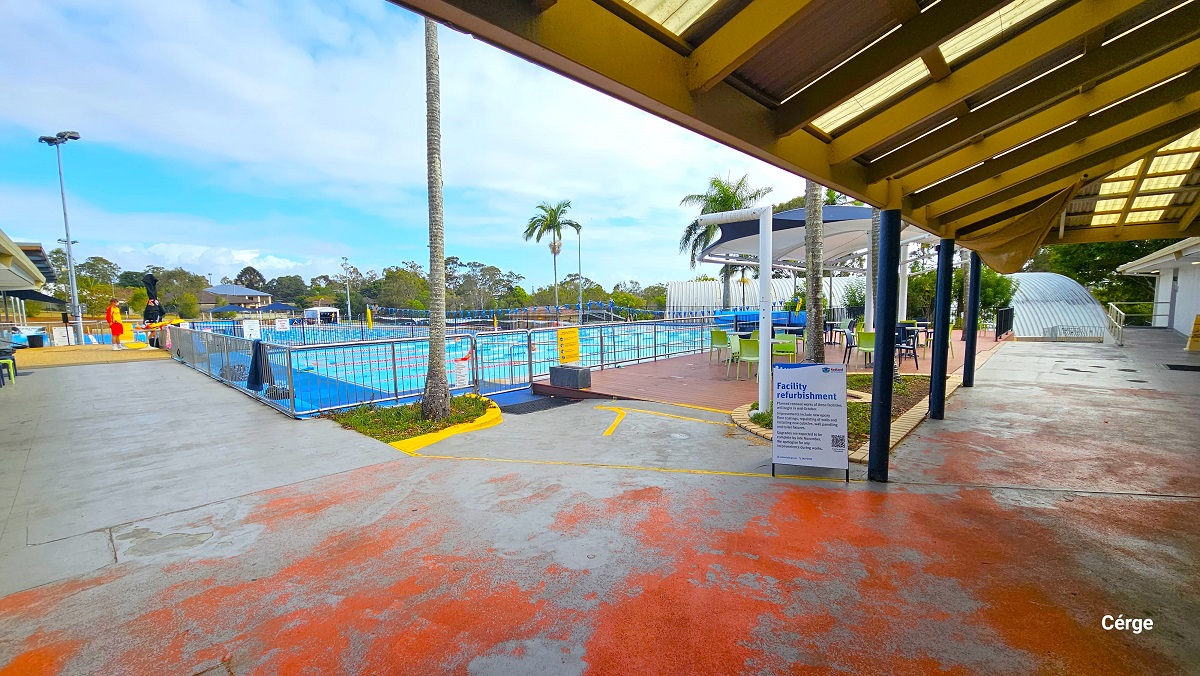 Outdoor aquatic areas at Cleveland Aquatic Centre. The outdoor pool is separated from the seating area on the right side by steel handrails placed in the entire pool perimeter. The seating section has lime green and blue plastic chairs with a white and blue overhang for shade. There are palm trees scattered within the outdoor area. This image also shows red markings on the floor.