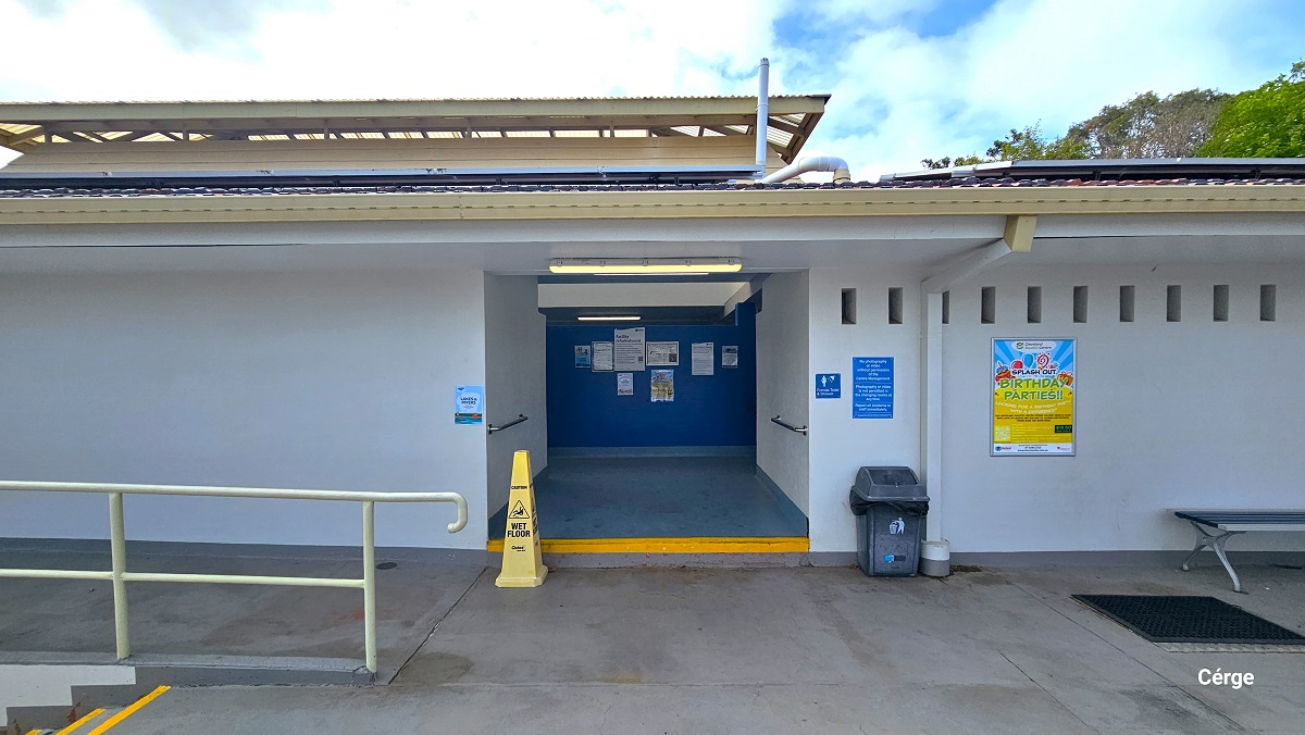 Access to the accessible bathrooms at Cleveland Aquatic Centre is through the stairs with cream-colored handrails and a ramp. On the bathroom entrance is a yellow cone hat for a wet floor. Against the right white wall is a grey garbage bin and a bench, while on the ground floor is a black mat. On the side wall of the smooth grey bathroom entryway are metal grab bars. Small adverts occupy the back blue wall.