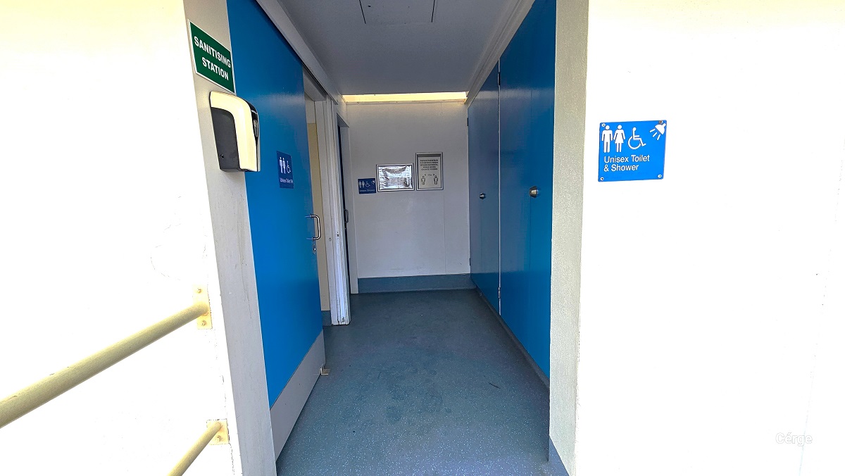 A closer look at the bathrooms at Cleveland Aquatic Centre. On the right white wall is the blue Unisex toilet and shower sign, while to the left white wall is the green sanitizing station label with a sanitizer dispenser. The bathroom doors are painted in blue and have a stainless steel door handle. The bathroom floor is grey.