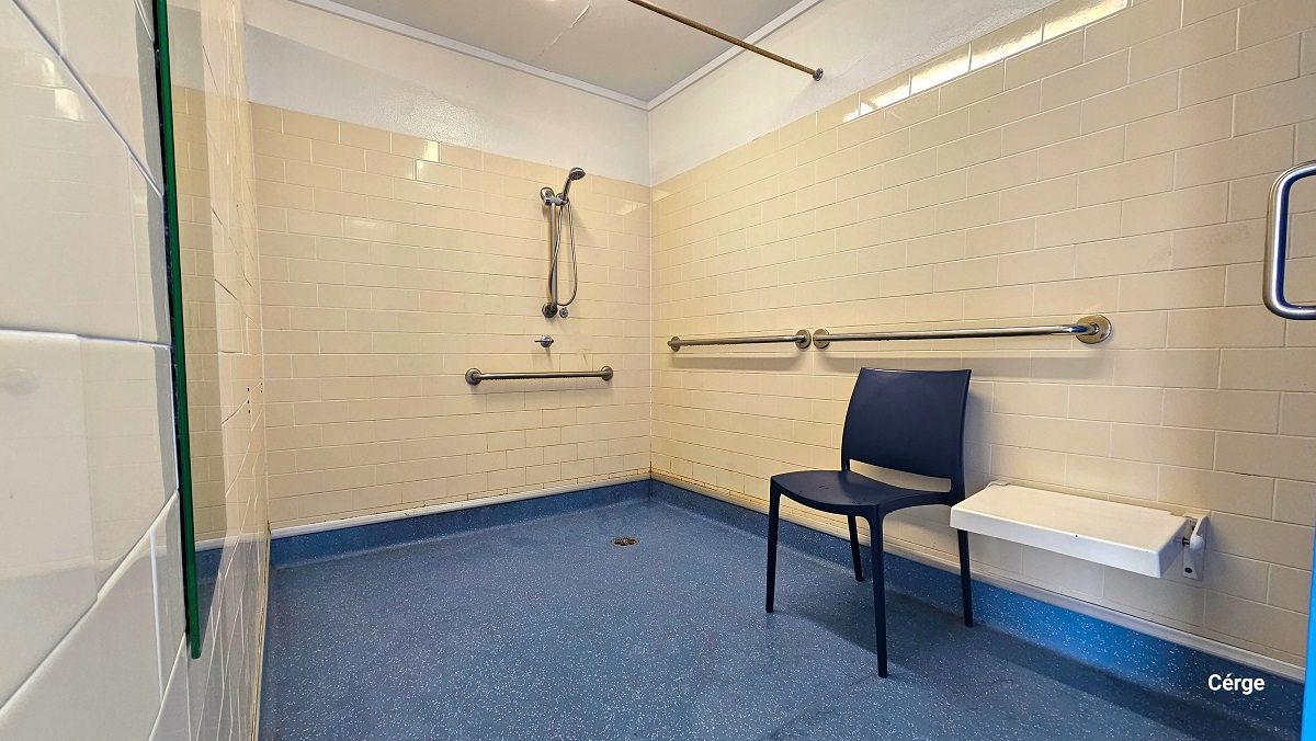 Accessible shower room at Cleveland Aquatic Centre. It has a blue with white dots floor, a white brick wall, and a white ceiling. Beside the wall-mounted white shower seat is a blue plastic chair. On the white brick walls with the shower head corner are grab bars to aid persons with mobility disabilities.