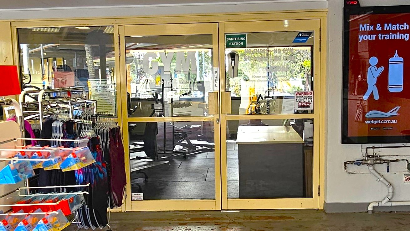 The glass entrance doors of the Gymnasium at Cleveland Aquatic Center have cream colored metal frames. It is manually operated as indicated by the word PULL in red font. On the right side there is a large red digital advert which advises guests to do a mix and match training. There is a sanitizing stating on the right door panel with a warning sign board that contains contact details. Right in front of the gym doors is a display of swimwear to the left.