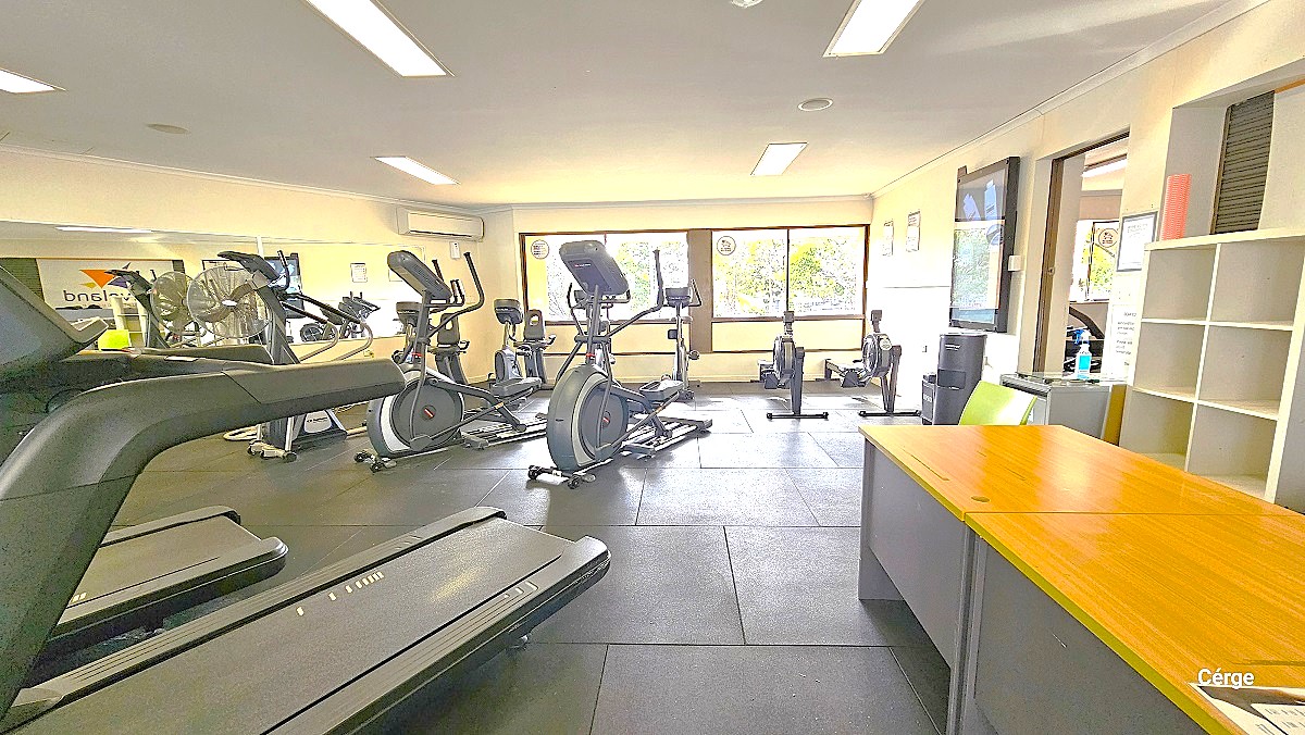 Image shows black cardio machines placed on top of the grey tile floor. The gym has a white ceiling with rectangular artificial ceiling fluorescent lights. A grey cabinet with polished wood top can be found on the right next to the white bag cabinet. The wall has glass windows that allow natural light inside the gym. The door on the right white wall connects to the free weights and resistance exercise machinery of the gym.