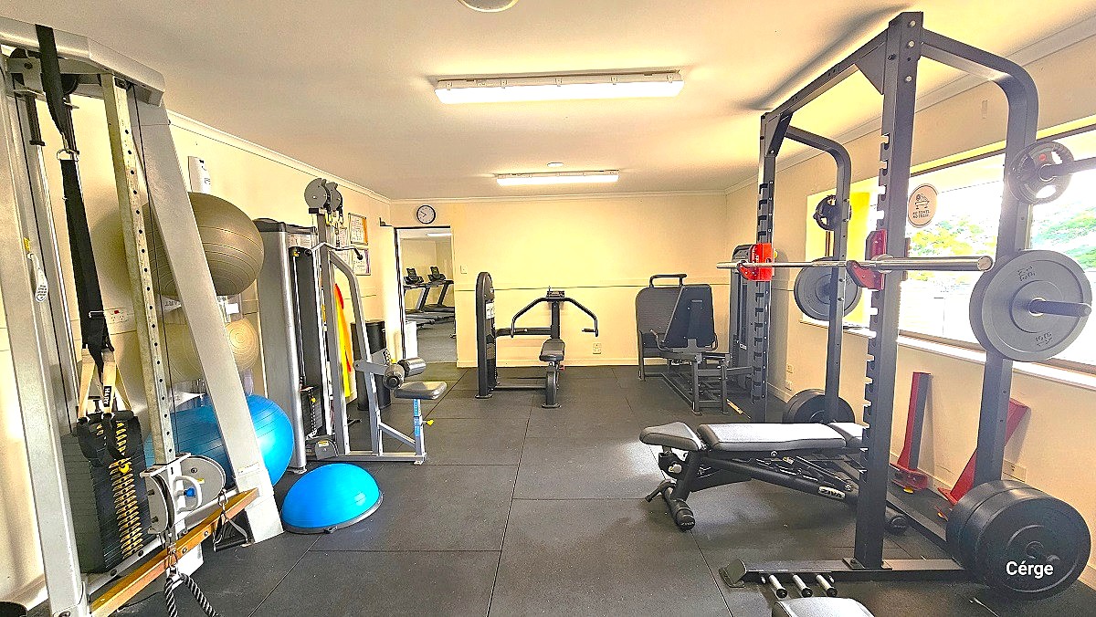 View opposite the entrance door of the free weights and resistance exercise machine. On the right side is a dumbbell rack with a black bench. On the left are brown, gold, blue and light blue exercise balls frequently used in Yoga. On top of the entrance door is a small clock.