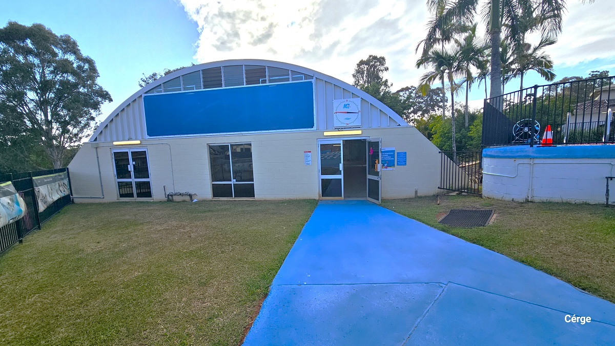 Entrance to the indoor pool at Cleveland Aquatic Centre. The indoor pool has a dome-like building structure and a combination of cream colored brick wall and a corrugated upper iron wall. It has three manually operated glass doors lined with white metal frames. The cemented pathway leading to the right door is painted in blue. At the upper part of the dome is a large blue rectangular shape. The perimeter is enclosed by as black vertical bar fence.