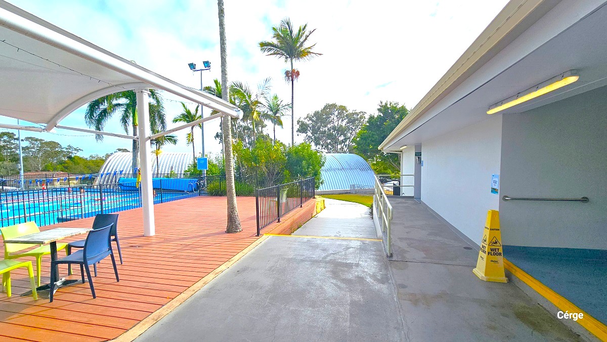 Smooth cemented pathway leading to the 20m indoor pool inside a long, corrugated iron domed structure is between the seating area near the 6 lane, 25m outdoor pool has a white dome cover with mahogany wooden floor and the bathrooms. It’s borders are the black vertical bar fence to the left and silver handrails to the right.