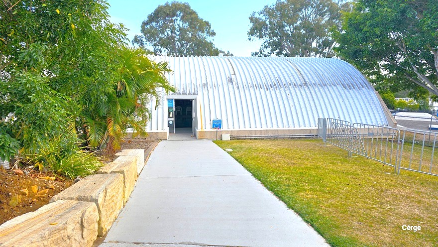 Smooth grey cemented pathway towards the entrance doors of the indoor pool at Cleveland Aquatic Centre. There is a grey fence on the right side with an open grass area. Rectangular rock boulders are lined on the garden to the left. The corrugated iron dome pattern of the indoor pool facility has some black markings on the surface. Gum trees and any types of plants thrive well as seen by the lush green leaves.