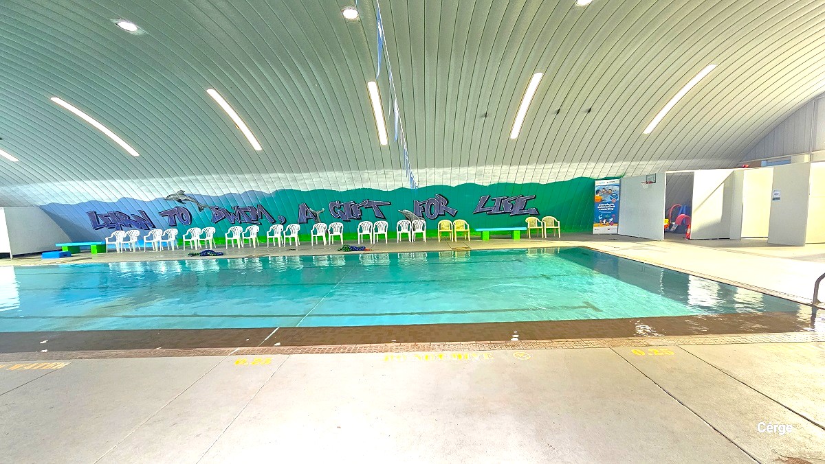 View of the indoor pool at Cleveland Aquatic Centre. The depth of the water is indicated next to the brown tiled pool wall which is 0.25 meter. A NO DIVE phrase is detailed in yellow also to the pool side. There is a blue bench and white plastic chairs on the opposite side of the pool, next to the green wall with graffiti that says, “Learn to Swim, A Gift for Life”. The ceiling has a corrugated iron roof dome pattern.