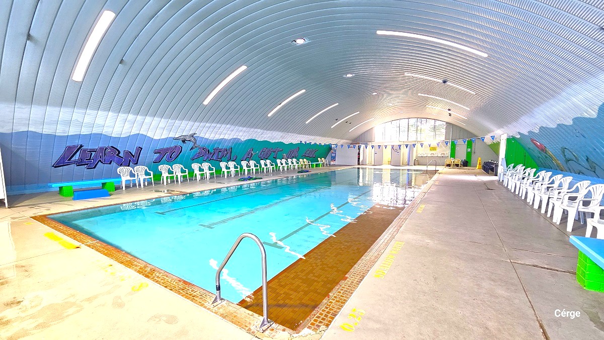 A large pool smooth pool deck is accessible around the indoor pool. The pool ladder can be distinguish by the inverted U shaped at the edge of the pool. Both the corrugated iron walls are painted with an ocean theme and a similar graffiti. White chairs with armrests and a bench are available on opposite sides of the pool. To the back is glass window that allows natural light to filter through and illuminate the facility. A string of blue triangular flags overhang the indoor pool.