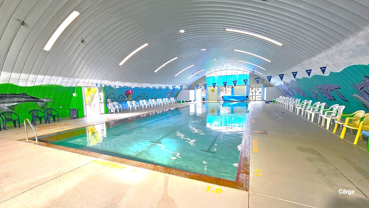 View of the indoor pool when standing near the blue triangular flags. The wall to the left is separated by the entrance door which has the Cerge app boy holding a QR code that aids persons with mobility disabilities and promotes inclusivity to every Center. Aside from the blue wall that imitates the ocean vibe, half of the wall is painted in green with a large fish. Place against this wall is a grey garbage bin and black plastic chairs.