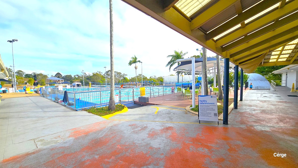 Outdoor aquatic area at Cleveland Aquatic Centre. The outdoor pool is separated from the seating area on the right side by steel handrails placed in the entire pool perimeter. The seating section has lime green and blue plastic chairs with a white and blue overhang for shade. There are palm trees scattered within the outdoor area. This image also shows red markings on the floor.
