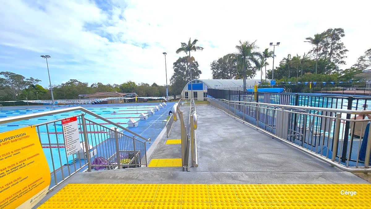 Entry to the 8 lane, 50m outdoor pool at Cleveland Aquatic Centre is via stairs and a ramp with handrails. Both are marked by yellow tiles with circular bumps at the entry point. On the left handrail are the pool rules details on yellow and white signboards. At the back is a white building with a dome roofing style.