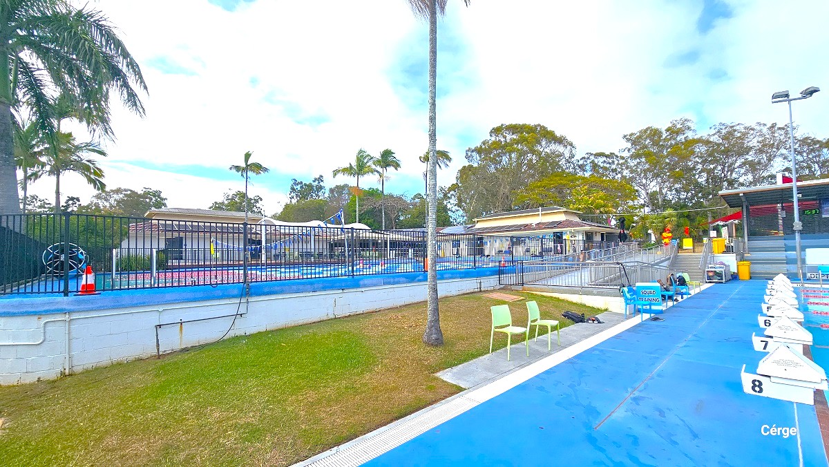 View from the pool deck of the 8 lane, 50m outdoor pool. Similar to 25m, 6 lane pool, the pool deck is painted in blue with white edges. There are two plastic lime-green chairs underneath a palm tree. A blue squad training signboard is placed in between blue plastic chairs found near the ramp entrance. At the start of the pool lanes are white diving blocks numbered from 1 to 8. To the back is the tiered grandstand overlooking the entire outdoor amenities.