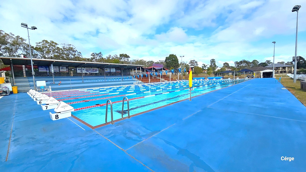 Right view of the 50m outdoor pool. The entire pool deck is blue. Each lane has a white diving block numbered from 1 to 8 and is separated by red, white, and blue floating rope lanes. There are pool ladders found on each side of the pool marked by the inverted U steel frame. Yellow garbage bins with cover can be found in front of the tiered grandstand to the back. Tall lamp posts are placed all throughout the surrounding area of the pool.