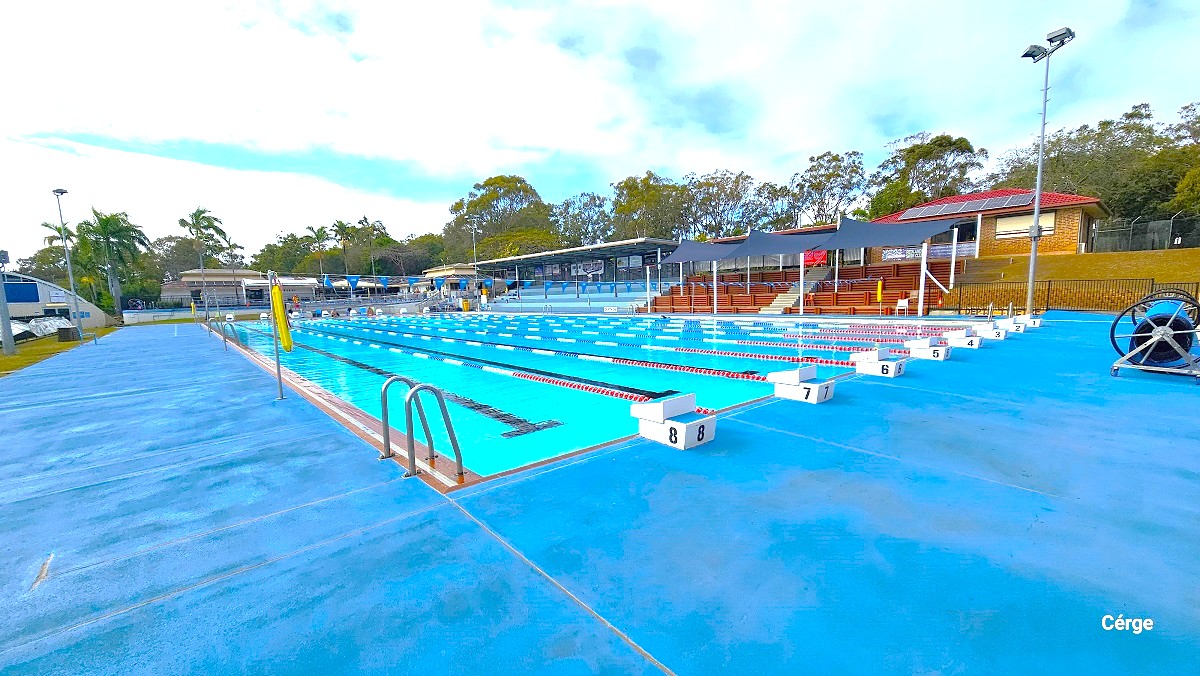 Cleveland Aquatic Centre has a 50m outdoor pool with a wide pool deck. On the right side is brown-tiered seating with a black sunshade and blue-tiered seating with a solid roof. Behind it are low-set brick cream-coloured buildings with brown tiled roofing having solar panels. Pool lanes are marked by white diving blocks marked from 1 to 8 and are separated by red, blue, and white floating rope lanes.