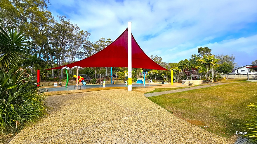 View after passing through the black vertical bar fence and near the yuca plants. The large red sunshade overhang at the top of the splash pool is wrapped around red, white, and yellow poles at three opposite ends of the pool. There is an adequate grass space on the right side. Different types of trees are planted all around the park.