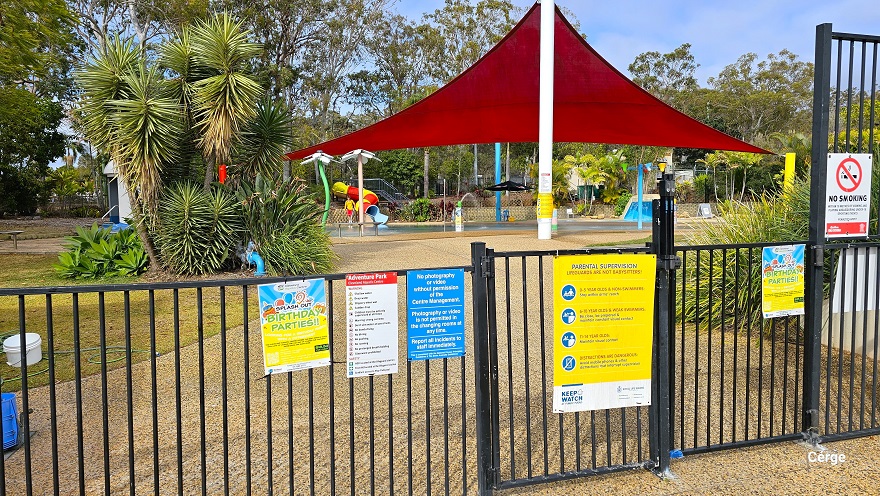 Splash park at Cleveland Aquatic Centre. Entry is through a low, black, vertical bar fence. Park rules and other information are detailed in the adverts hanging on the fence. There is a no smoking sign on the upper right of the adult high vertical bar fence. The natural stone tile pathway, passing through the yuca plant on the left side, leads to the splash pool. There is a large red triangular sunshade that offers shade to the splash pool.