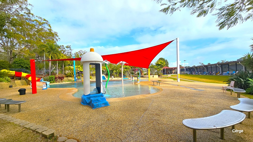 The pool deck floors are made of natural stone flooring. Near the edges are silver metal, slightly curved benches. There are tall, green, and white spraying fountains with a white flower petal-like top. On the left of the pool is a spiraling, red, and yellow water slide. Covering the perimeter of Splash Park is a corrugated wall seen further right with white tube lining.