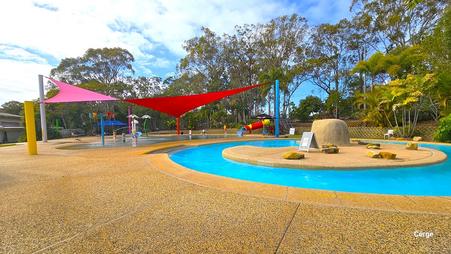 Adventure pool next to the Splash Park at Cleveland Aquatic Centre. The pool has a moon-shaped center with a large rock at the center. There were also small rocks on the floor of the moon-shaped centerpiece. From this position, there is a pink triangular sunshade next to the red sunshade. A stone wall separates the pool deck and garden area.