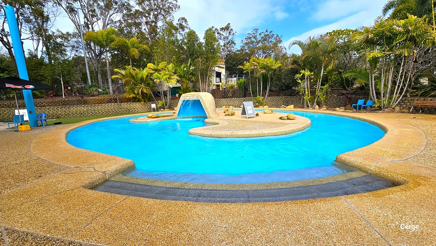A closer look at the Adventure pool at Cleveland Aquatic Centre. The pool has a bell shape with a moon-shaped center. A large stone at the center is hollow inside for a waterfall effect. Underneath the palm trees against the stone wall is a wooden bench and blue and white plastic chairs. Next to the blue pool is a black Pepsi sunshade with two blue monoblock chairs.