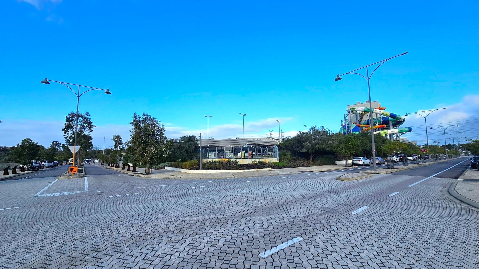 A wide, paved road with a textured surface runs through the foreground. On the left, there are trees lining the street and parked cars. In the centre, a building with a modern design is visible, surrounded by greenery. On the right, a colourful water slide structure is prominent, with more parked cars and trees. Tall streetlights line both sides of the road.