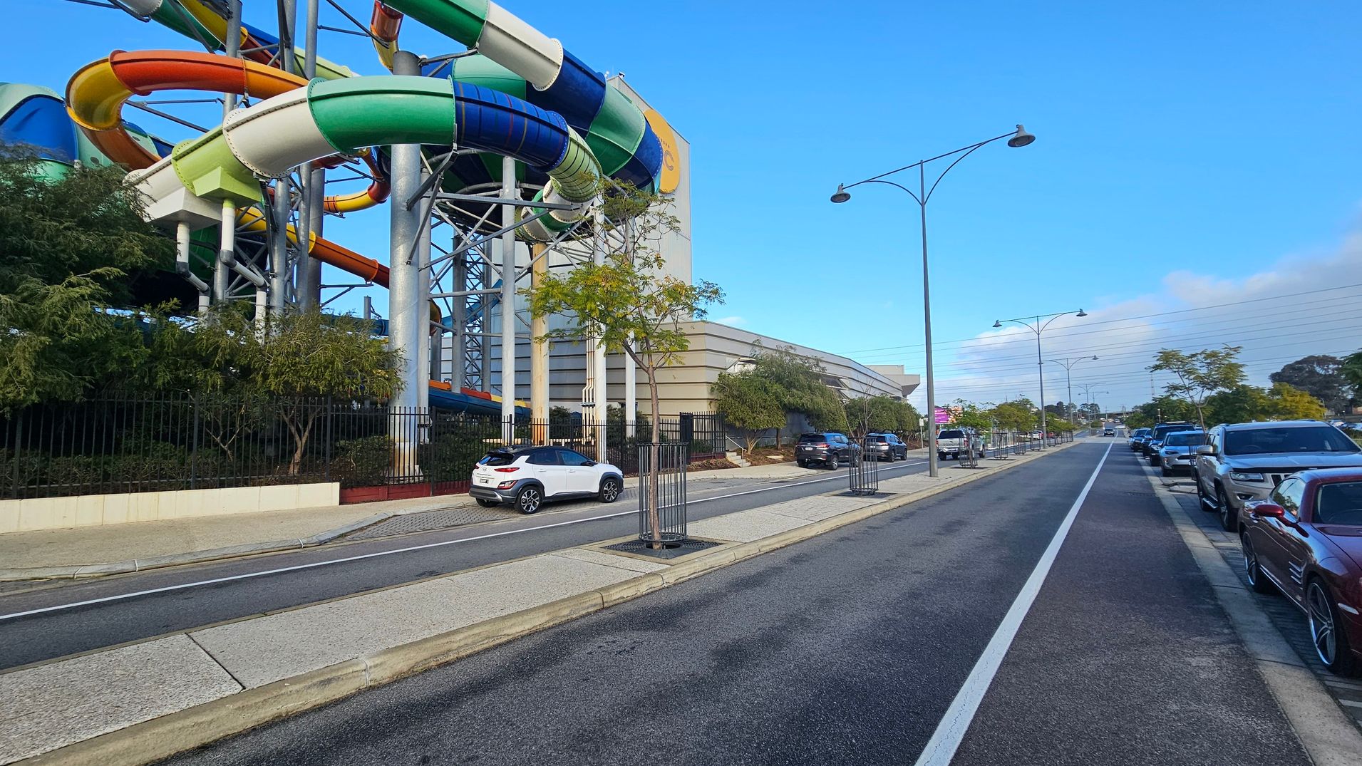 On the left, there are colourful water slides supported by metal structures, surrounded by trees and a black fence. A white car is parked on the adjacent road. On the right, a street lined with parked cars extends into the distance, bordered by trees and street lamps. The road has a marked bike lane. A large building with horizontal lines is visible in the background. The pavement is light grey, and the road is dark grey.