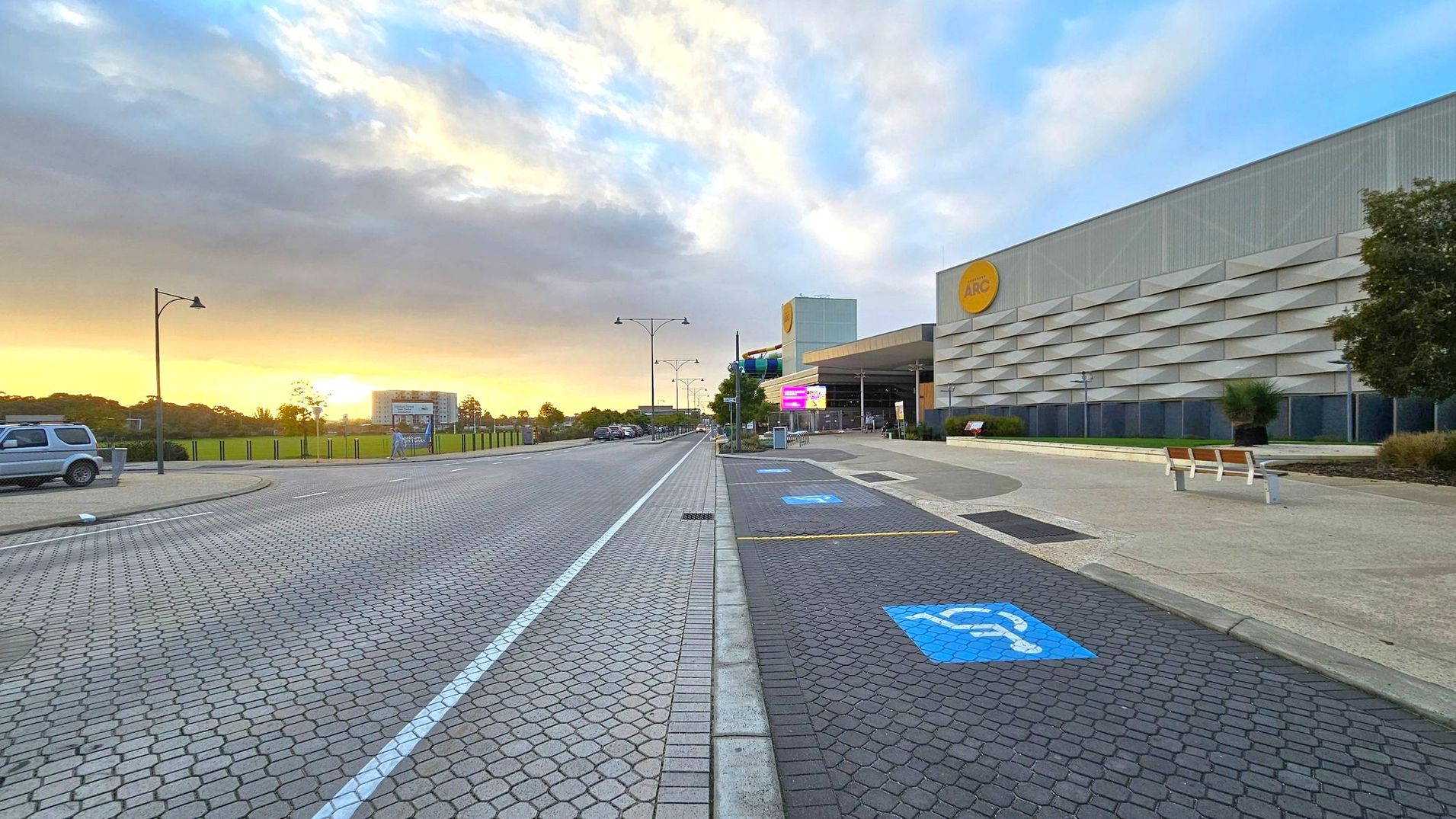 A paved road with a cobblestone pattern runs through the centre, flanked by a pavement on the right. On the right, a large building with a grey facade and a yellow circular logo is visible. There are designated parking spaces with blue markings, including disabled parking. Benches and a tree are also present. On the left, a grassy area with a fence and a parked vehicle can be seen. Street lamps line the road, and the sky is partly cloudy with a warm glow near the horizon.