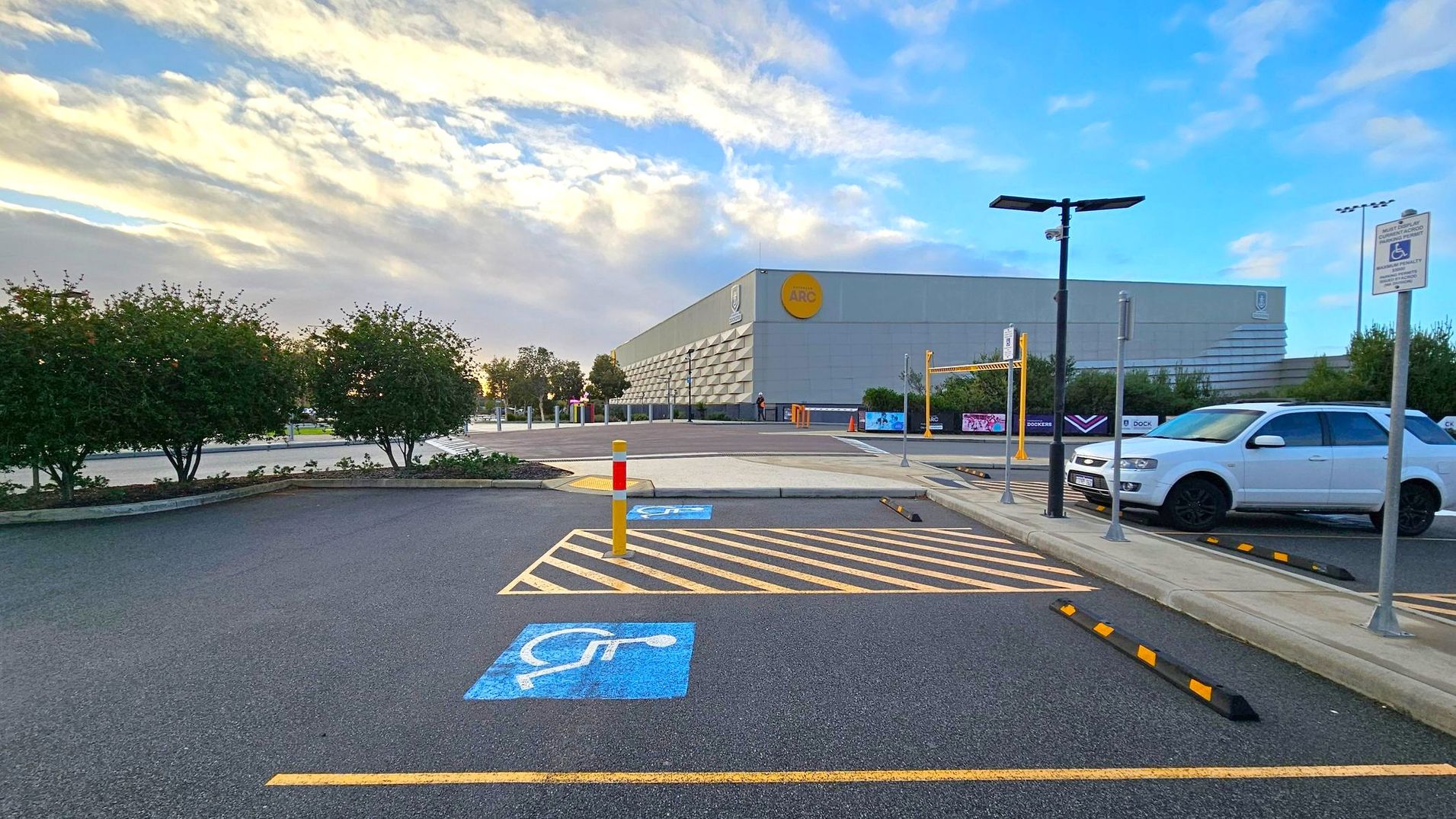 A car park with a designated disabled parking space marked in blue on the asphalt. On the right, a white SUV is parked next to a sign indicating the disabled parking area. In the background, a large grey building with a yellow circular logo is visible. There are several trees and bushes lining the perimeter of the car park. A tall streetlight and a barrier gate are also present near the building. The sky is partly cloudy.