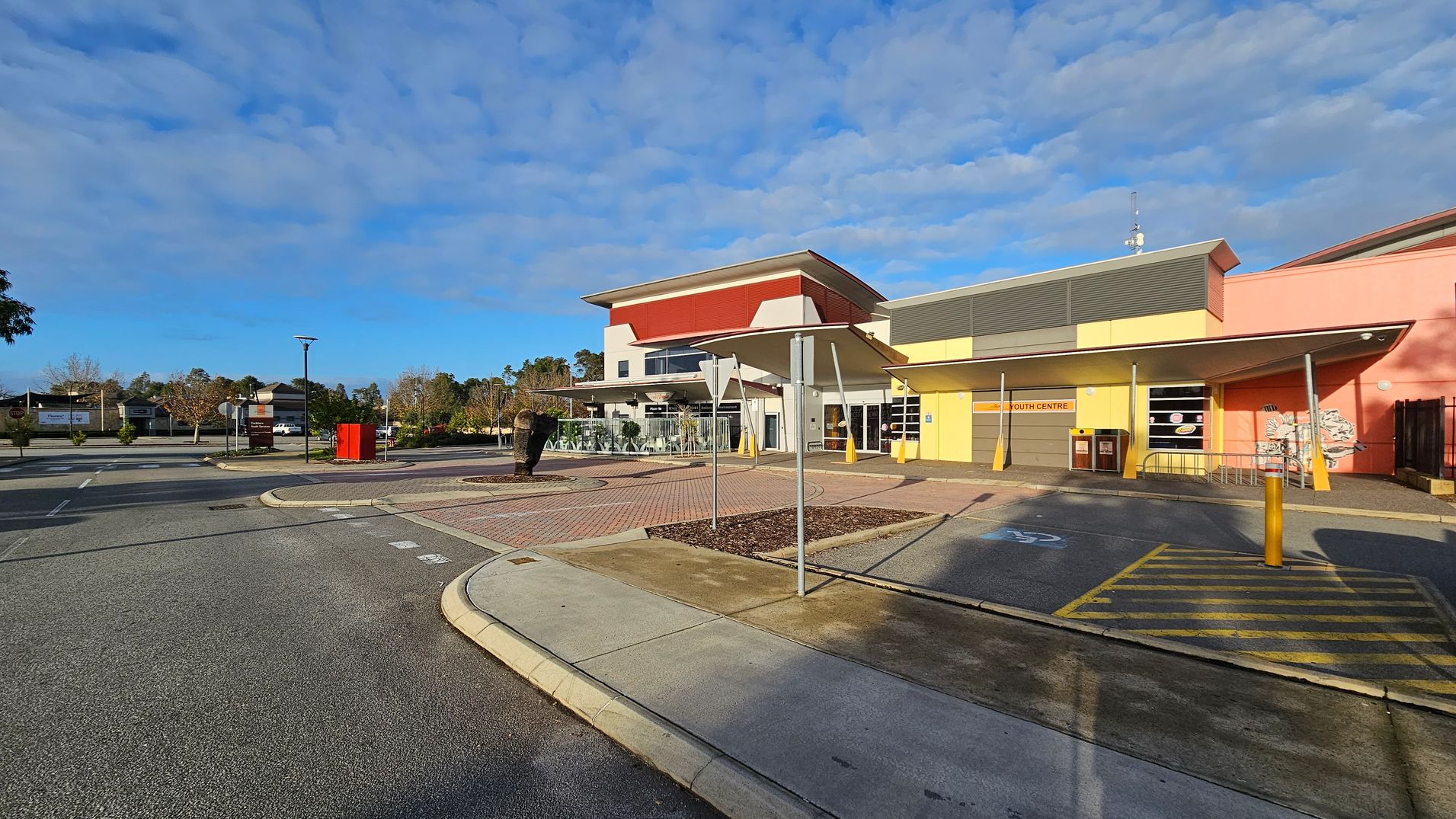 A modern building with a red and white facade is on the left, featuring large windows and a shaded outdoor area. To the right, a yellow and pink structure labelled "Youth Centre" has a sloped roof and several windows. In the foreground, a paved area with a roundabout and a sculpture is visible, along with a pedestrian crossing marked in yellow. The background includes trees and a few parked cars. The ground is a mix of concrete and asphalt.