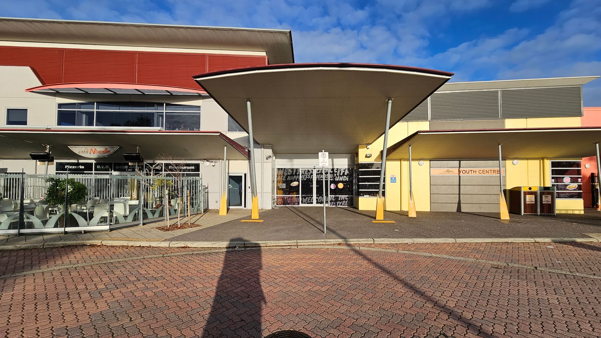 In the foreground, a paved area with a shadow cast on it. The middle features a building with a central entrance under a large, curved canopy supported by poles. To the left, a café with outdoor seating enclosed by a metal fence. To the right, a section marked "Youth Centre" with a yellow facade and two bins. The building has a mix of red, yellow, and grey panels, with windows and signage.