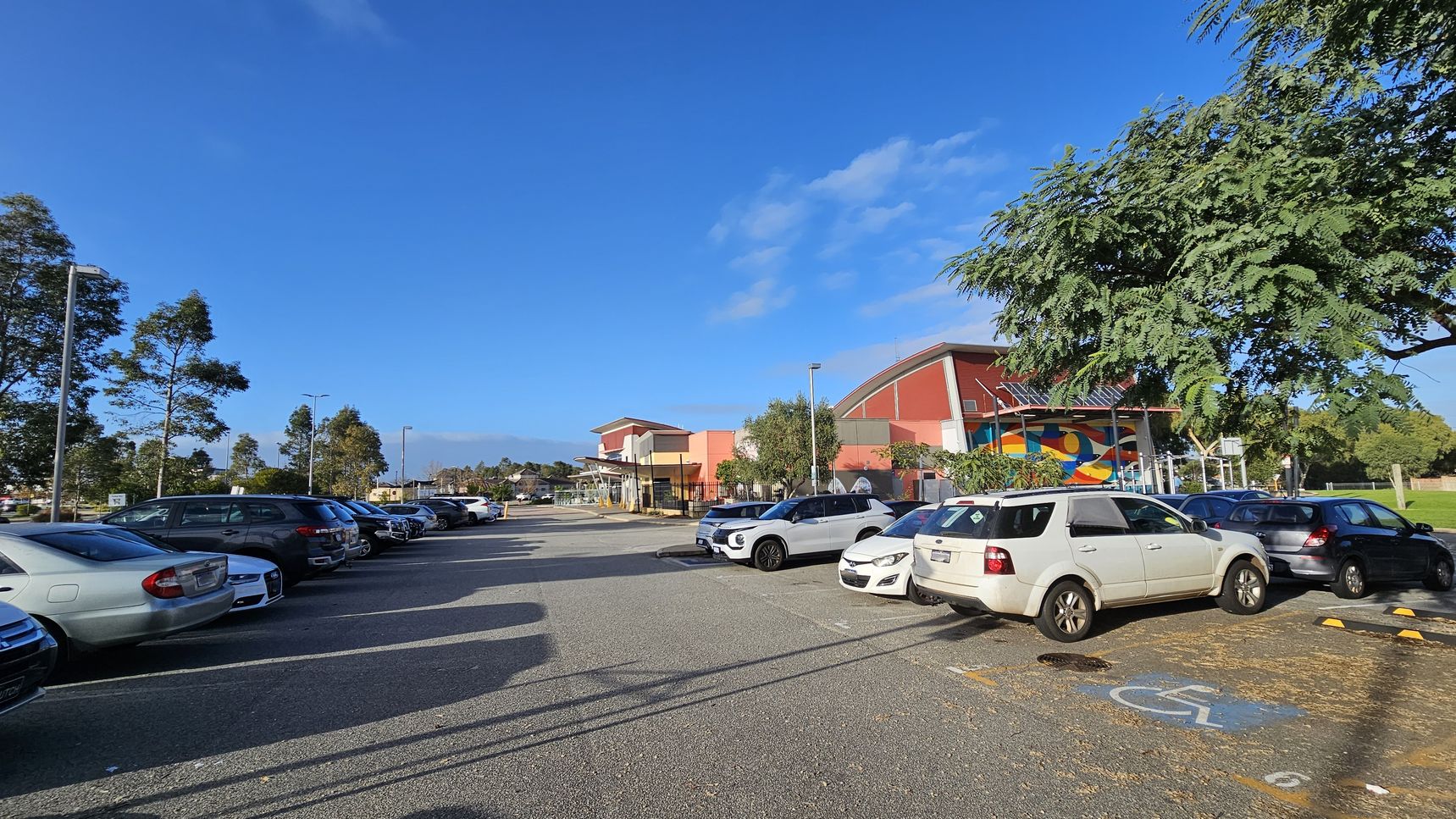 A car park with several vehicles is on the left, including a white SUV in the foreground. On the right, a building with a colourful mural and a red roof is partially visible, surrounded by trees. The ground is paved, and there is a marked disabled parking space in the foreground. Tall streetlights and more trees are in the background under a clear blue sky.