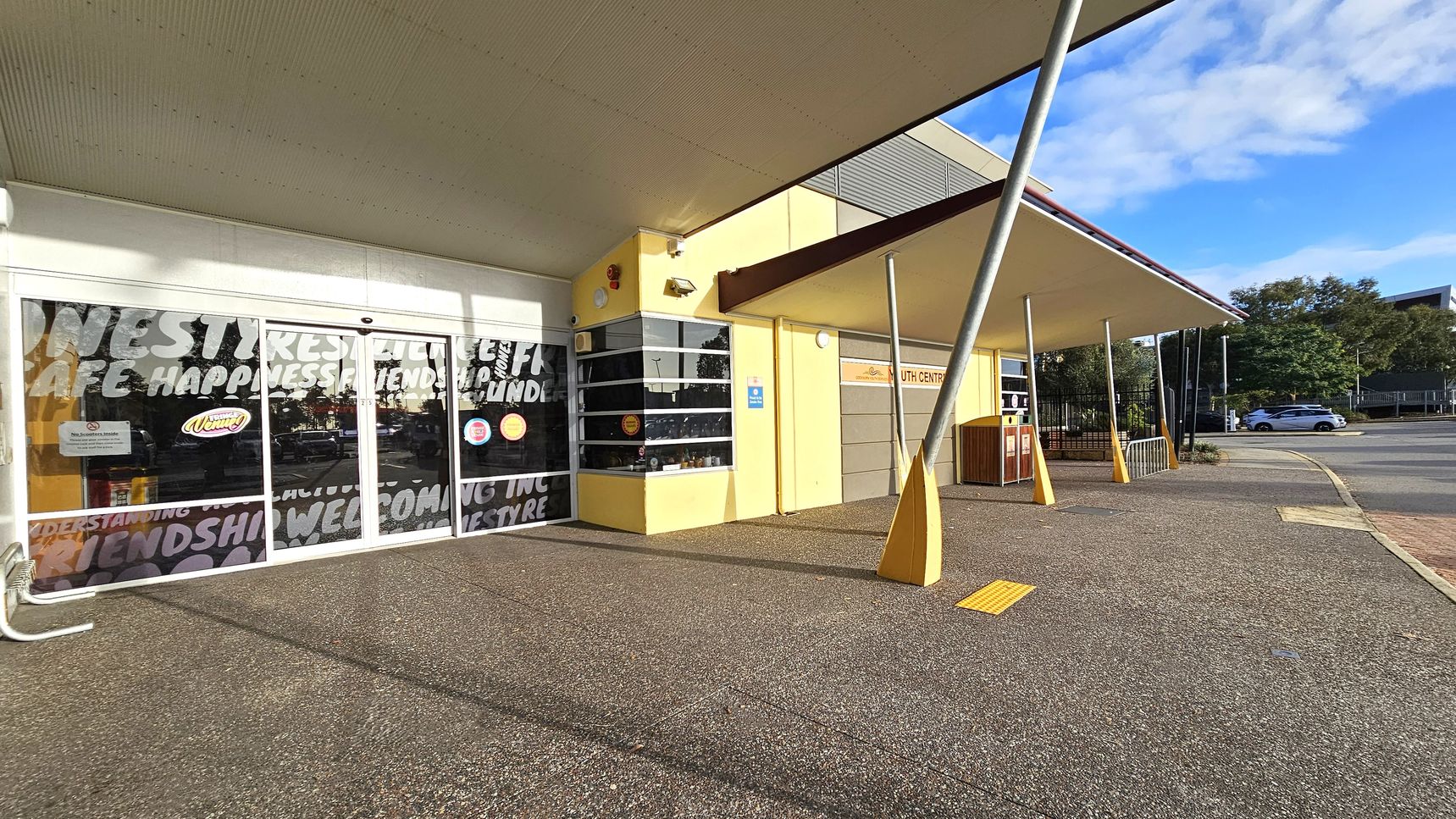 A building with a yellow exterior and a large overhanging roof. On the left, there are glass doors with words like "happiness" and "friendship" printed on them. The right side features a sign for a youth centre and a series of angled metal poles supporting the roof. The ground is paved with a textured surface, and a yellow tactile paving strip is visible. In the background, there are trees and a parked car on the street.