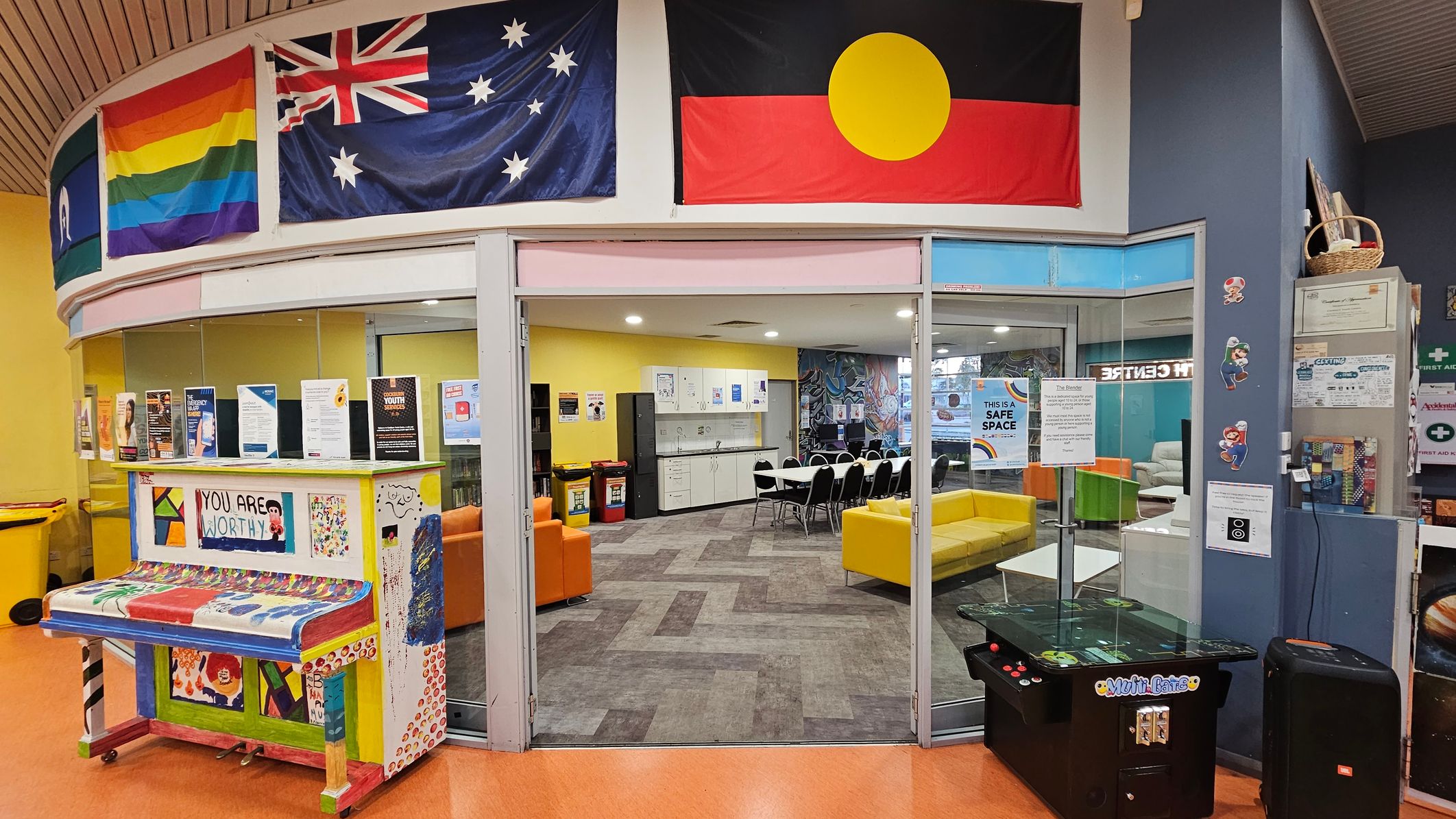 A colourful indoor space with a curved ceiling. On the left, a vibrant piano with "YOU ARE WORTHY" painted on it is surrounded by various posters. Above, flags including a rainbow flag and the Australian flag are displayed. In the centre, a glass entrance leads to a room with yellow and orange seating, a table with chairs, and a kitchenette. On the right, a gaming machine and wall decorations featuring characters are visible. The floor is a mix of orange and patterned tiles.