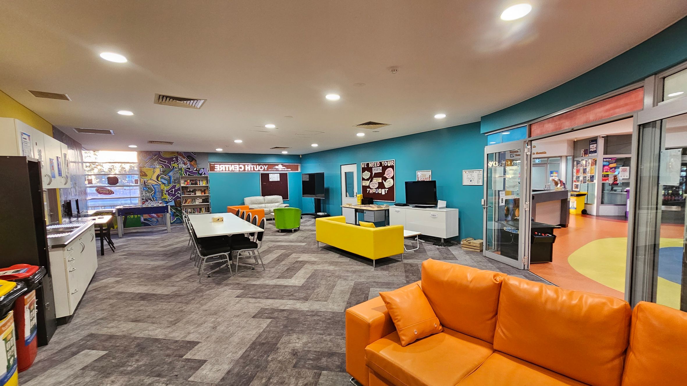 A spacious room with a vibrant, modern design. On the left, there are white cabinets and a row of computers. In the centre, a long table with black chairs is surrounded by colourful seating, including a yellow sofa and an orange couch. The back wall features a graffiti-style mural and bookshelves. On the right, glass doors lead to another area with an orange and yellow floor. The ceiling is fitted with recessed lighting, and the floor has a grey, patterned design.