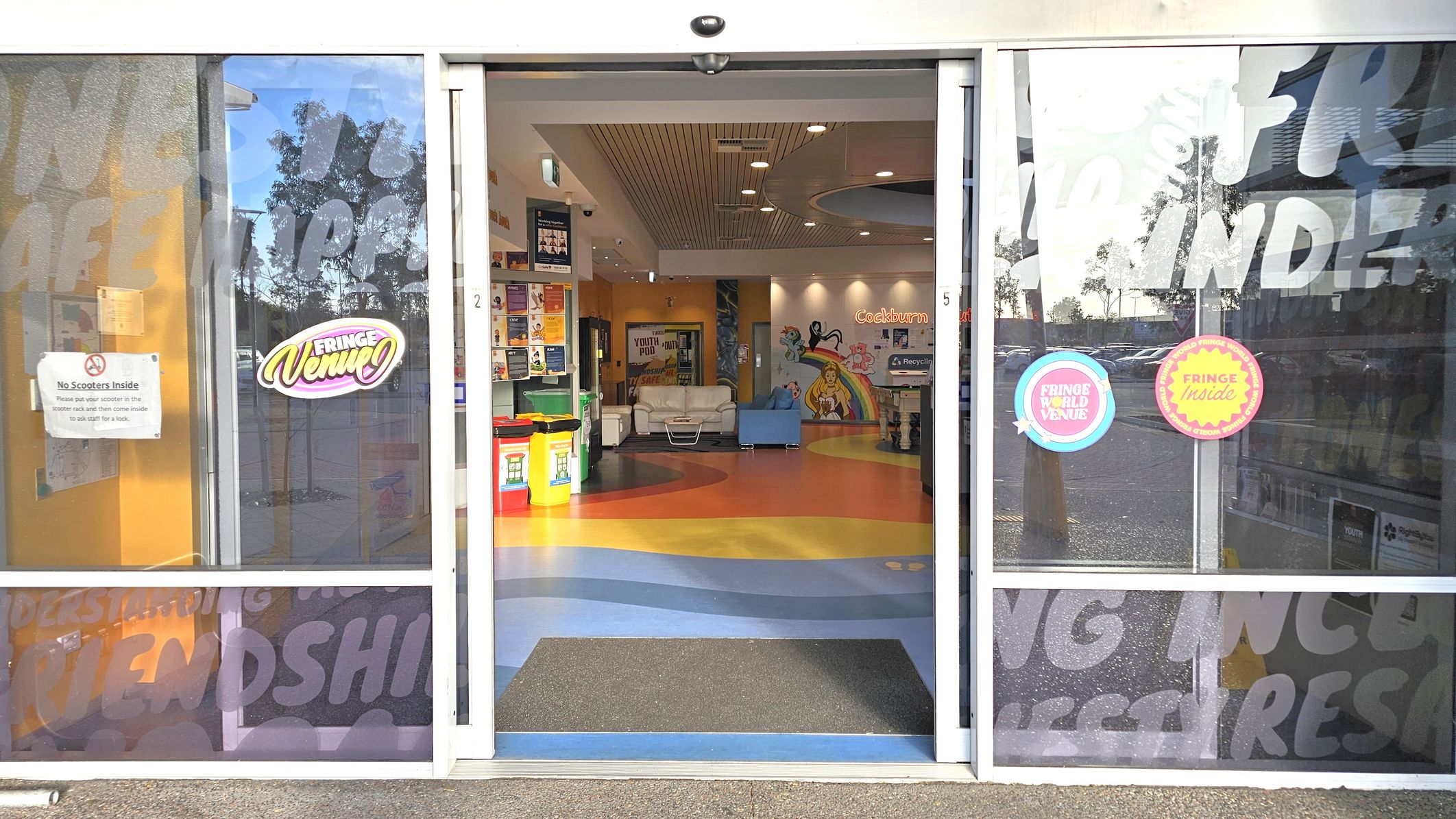 Sliding glass doors with signage, including "Fringe Venue" and "No Scooters Inside". Inside, a colourful floor with blue, yellow, and orange patterns leads to a seating area with white and blue couches. Walls are decorated with vibrant murals and posters. A recycling bin is visible on the right. The ceiling has recessed lighting and a wooden panel design. Reflections of trees and parked cars are seen on the glass.