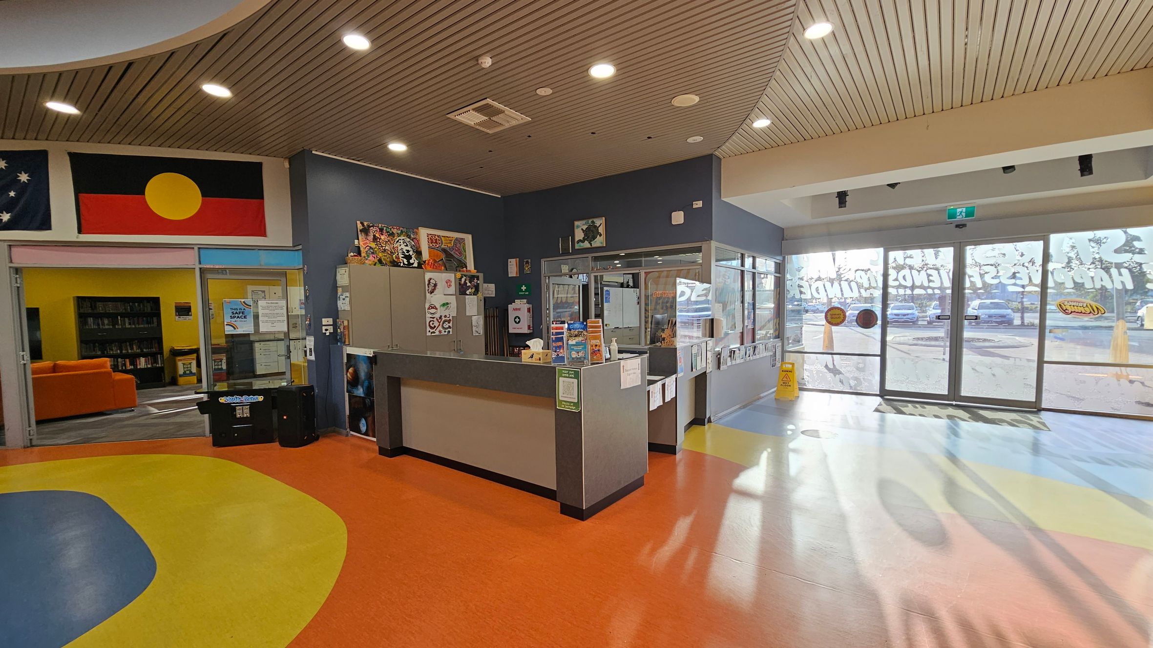 A reception area with a colourful floor featuring orange, yellow, and blue patterns. On the left, there is a library entrance with an orange sofa and bookshelves visible through glass doors. Above, flags are displayed on the wall. In the centre, a reception desk is adorned with various notices and items. To the right, large glass doors lead outside, with a wet floor sign nearby. The ceiling is wooden with recessed lighting.