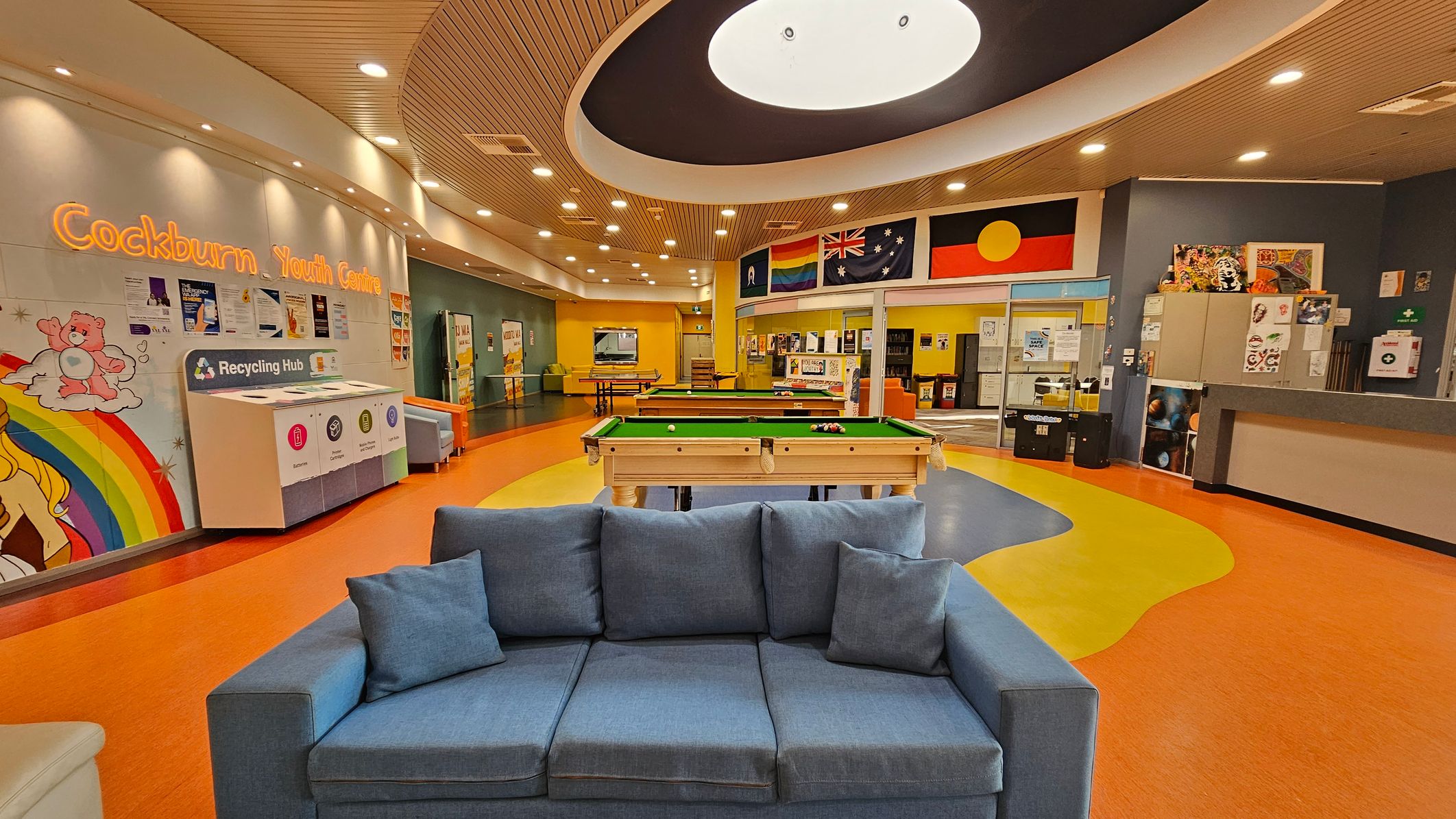 A vibrant indoor space with a blue sofa in the foreground. In the middle, there are pool tables on a colourful floor with orange, yellow, and blue patterns. The background features a wall with flags and a sign reading "Cockburn Youth Centre." On the left, there's a recycling hub and a mural with a rainbow and cartoon characters. The ceiling is curved with recessed lighting and a large circular light fixture.