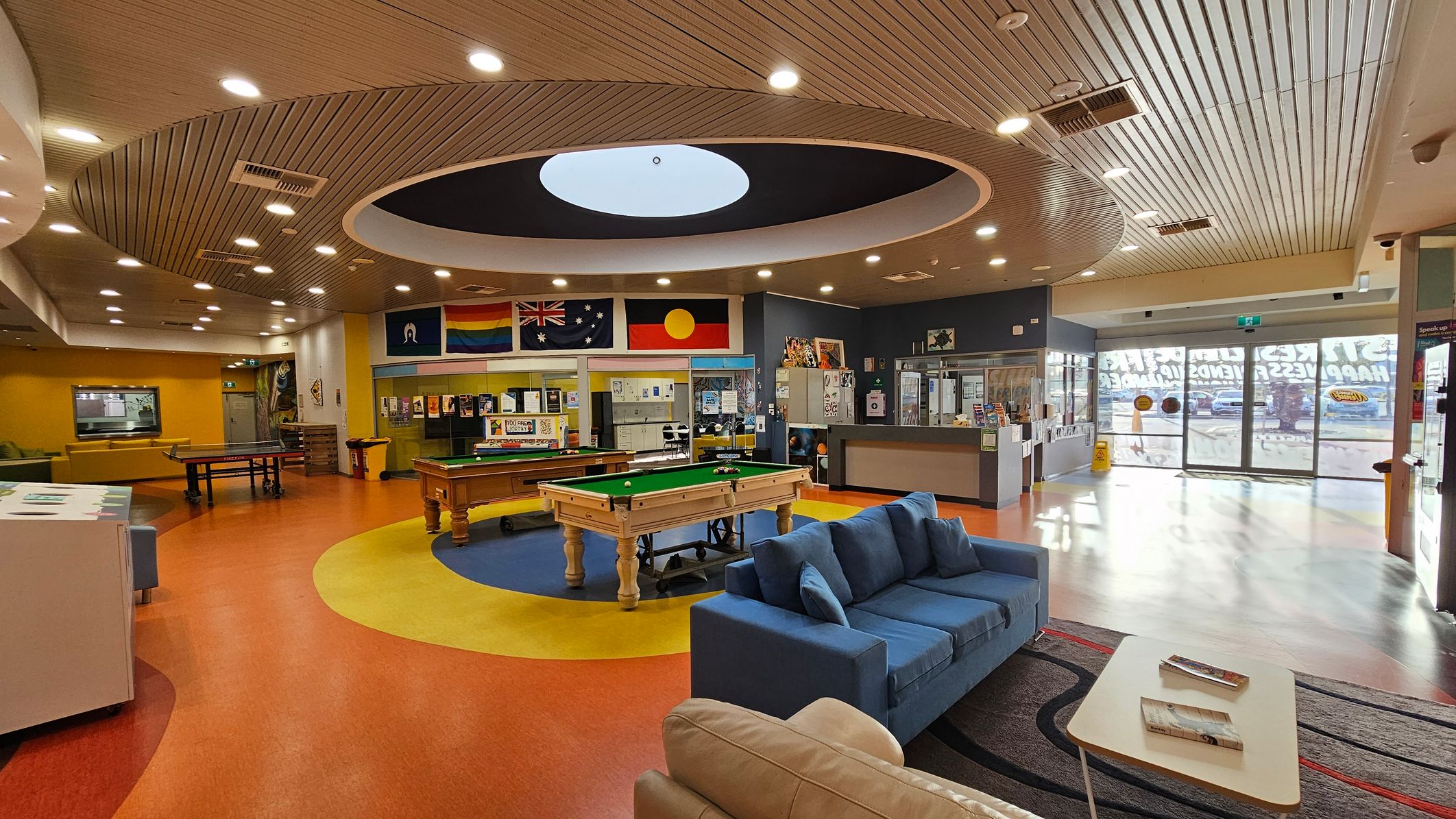 A spacious indoor recreational area with a circular ceiling design featuring recessed lighting. In the centre, there's a pool table on a blue and yellow circular carpet. To the left, a ping pong table and yellow seating area. On the right, a blue sofa and a small table with books. The back wall displays various flags and posters. The floor is a mix of red, yellow, and blue, and the entrance has large glass doors with signage.