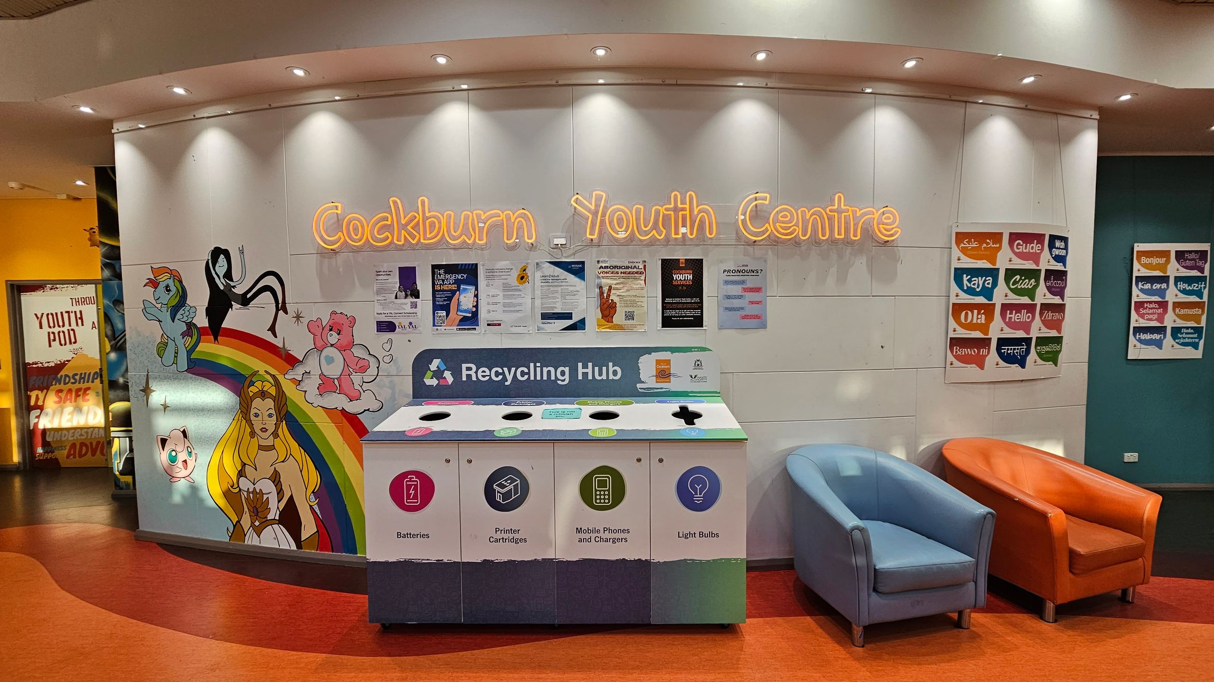 A colourful wall with the words "Cockburn Youth Centre" in neon lights. Below, a recycling hub with sections for batteries, printer cartridges, mobile phones, and light bulbs. To the left, a mural featuring cartoon characters and a rainbow. On the right, two chairs, one blue and one orange, sit on an orange floor. Posters with greetings in various languages are displayed on the right wall. The ceiling has recessed lighting.