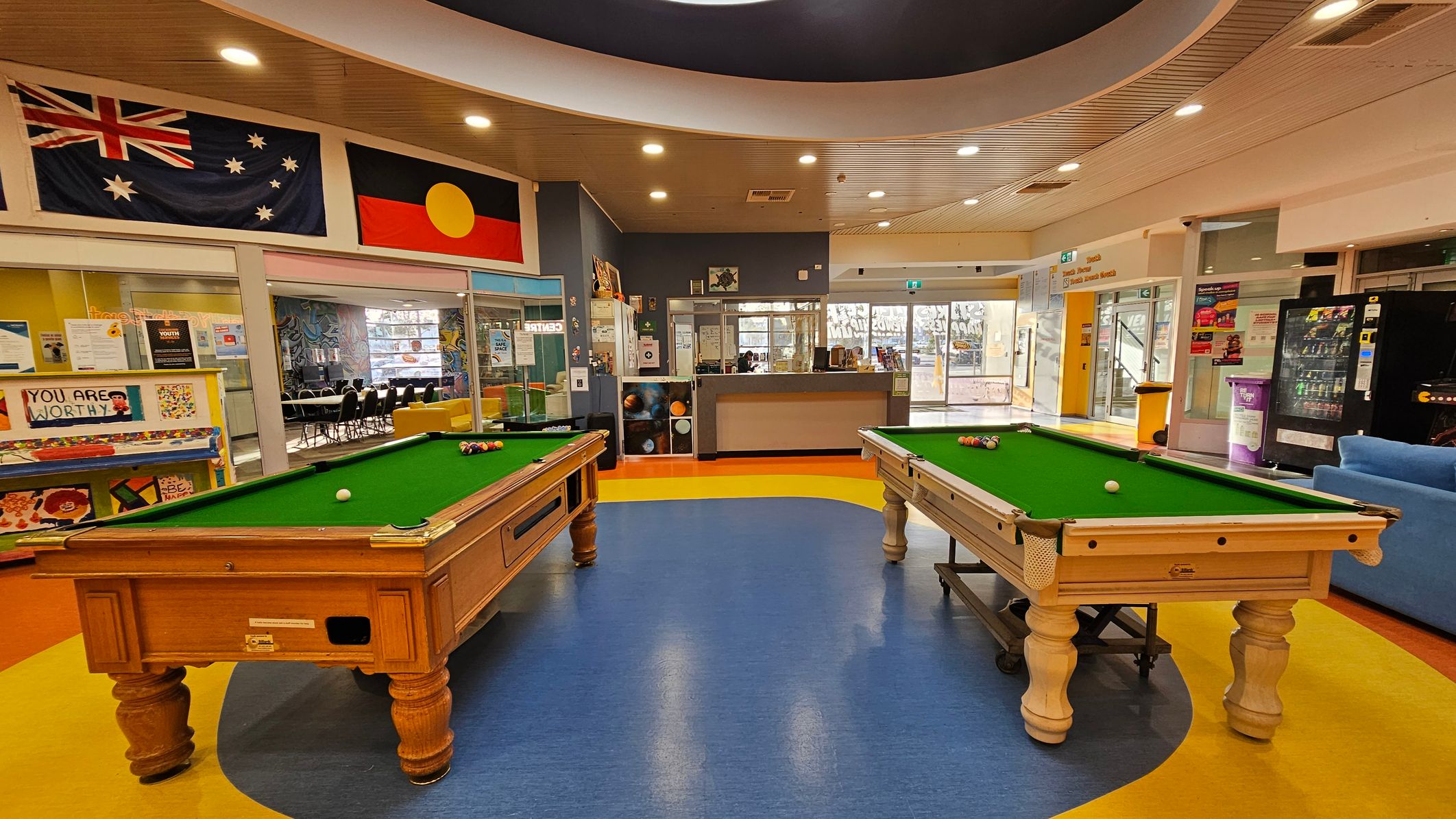 Two pool tables with green felt tops are positioned in the centre of a room with a blue and yellow floor. Above, the ceiling is curved with recessed lighting. On the left wall, there are Australian and Aboriginal flags. Behind the pool tables, a glass partition reveals a room with chairs and tables. To the right, a vending machine and a blue sofa are visible. The background includes a reception area with a counter and glass doors leading outside.