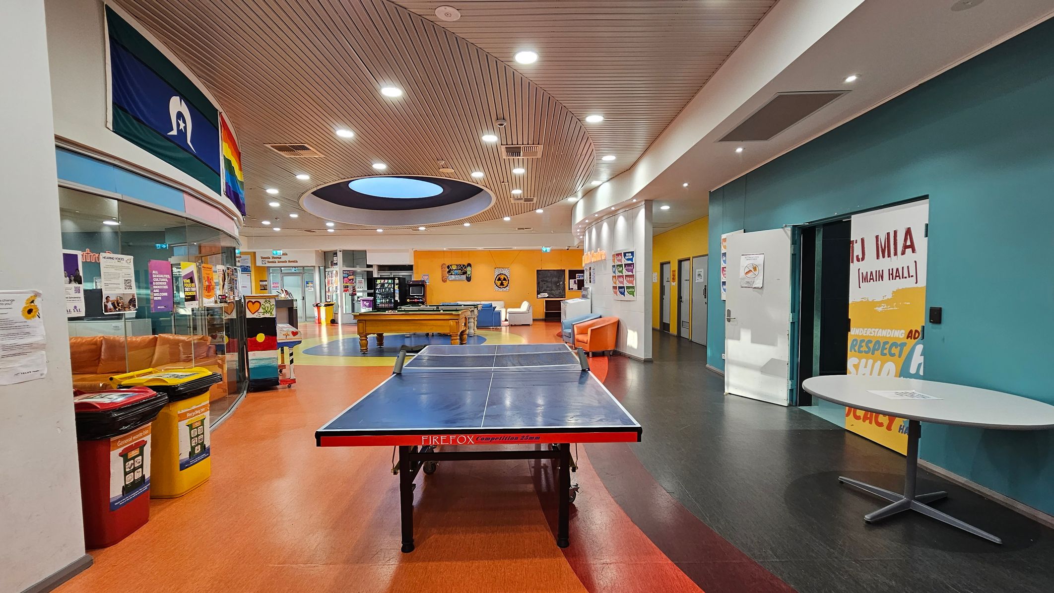 A spacious indoor recreational area with a ping pong table in the centre on an orange floor. To the left, there are colourful bins and a curved glass wall with posters and a rainbow flag. The right side features a teal wall with a door labelled "Main Hall" and a round table. In the background, there's a pool table, vending machines, and seating areas with couches. The ceiling is curved with recessed lighting and a circular skylight.