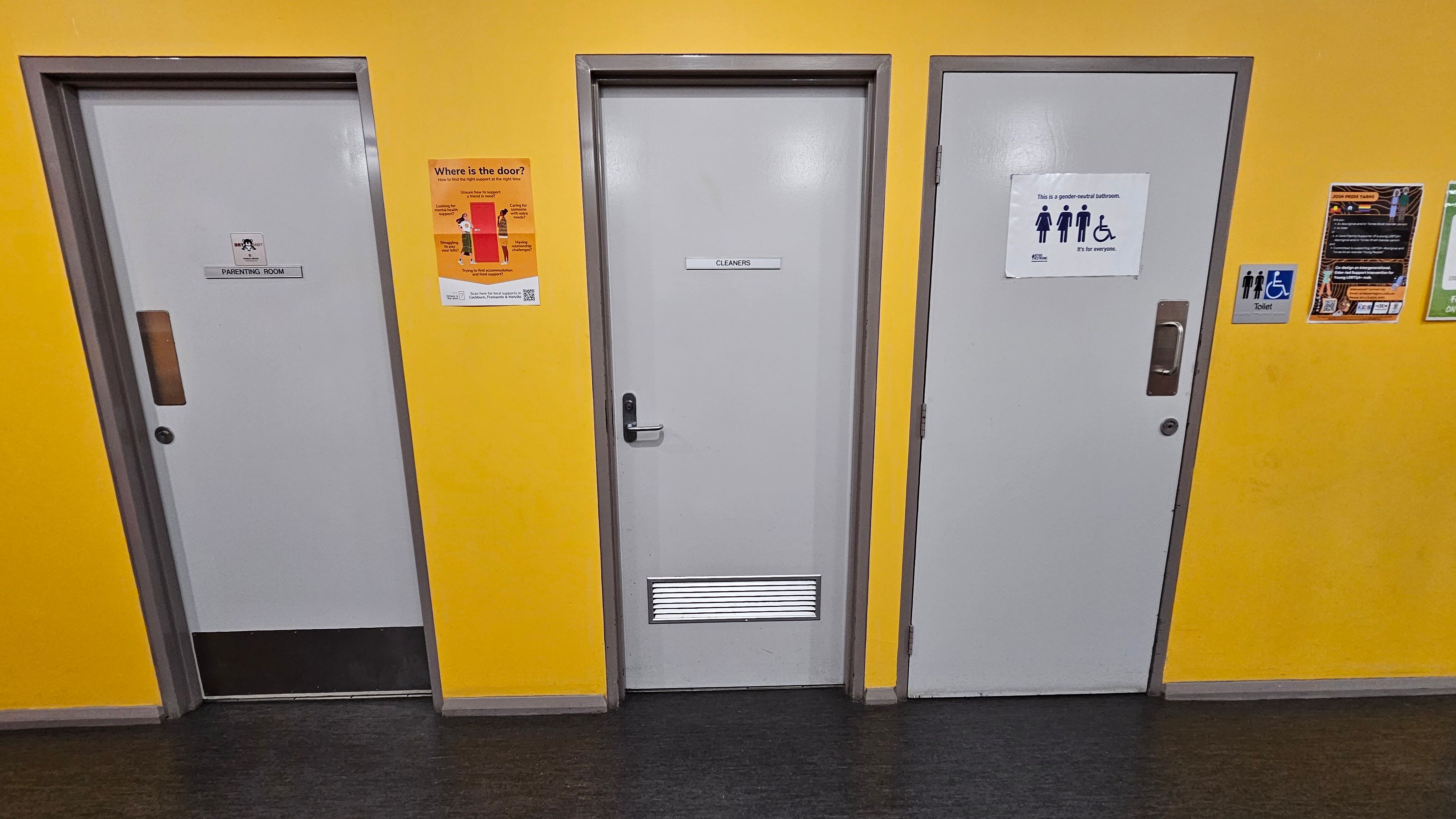 Three doors are set in a bright yellow wall. The left door is labelled "Parenting Room" with a small window. The middle door is marked "Cleaners" and has a vent at the bottom. The right door is a gender-neutral bathroom, indicated by a sign with symbols for male, female, and wheelchair access. Various informational posters are on the wall, including one about door locations. The floor is dark grey, and the ceiling is not visible.