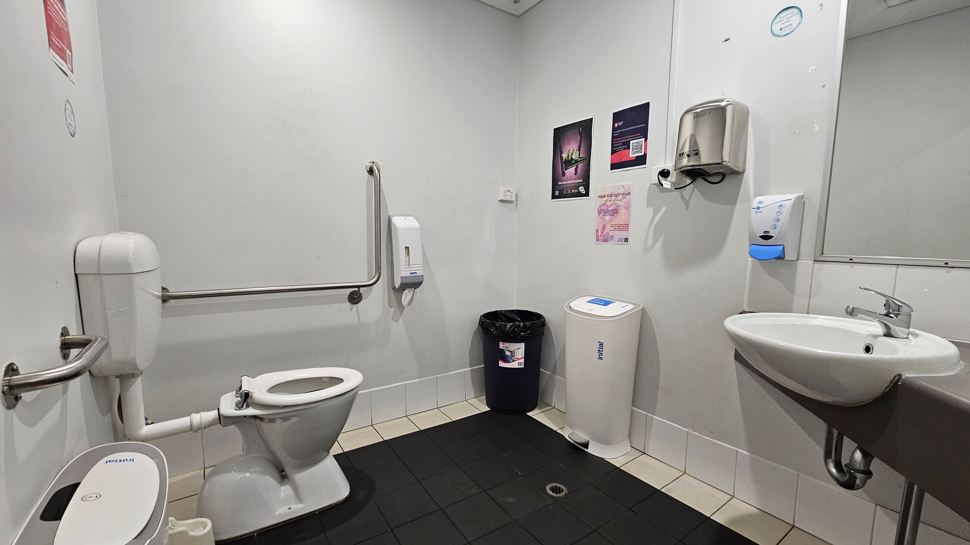 A public restroom with a white toilet on the left, equipped with a support rail. A black bin and a white sanitary bin are against the back wall. The right side features a wall-mounted sink with a silver tap, a soap dispenser, and a hand dryer above. The walls are adorned with posters. The floor is tiled in black and beige, and the ceiling is plain white with recessed lighting.