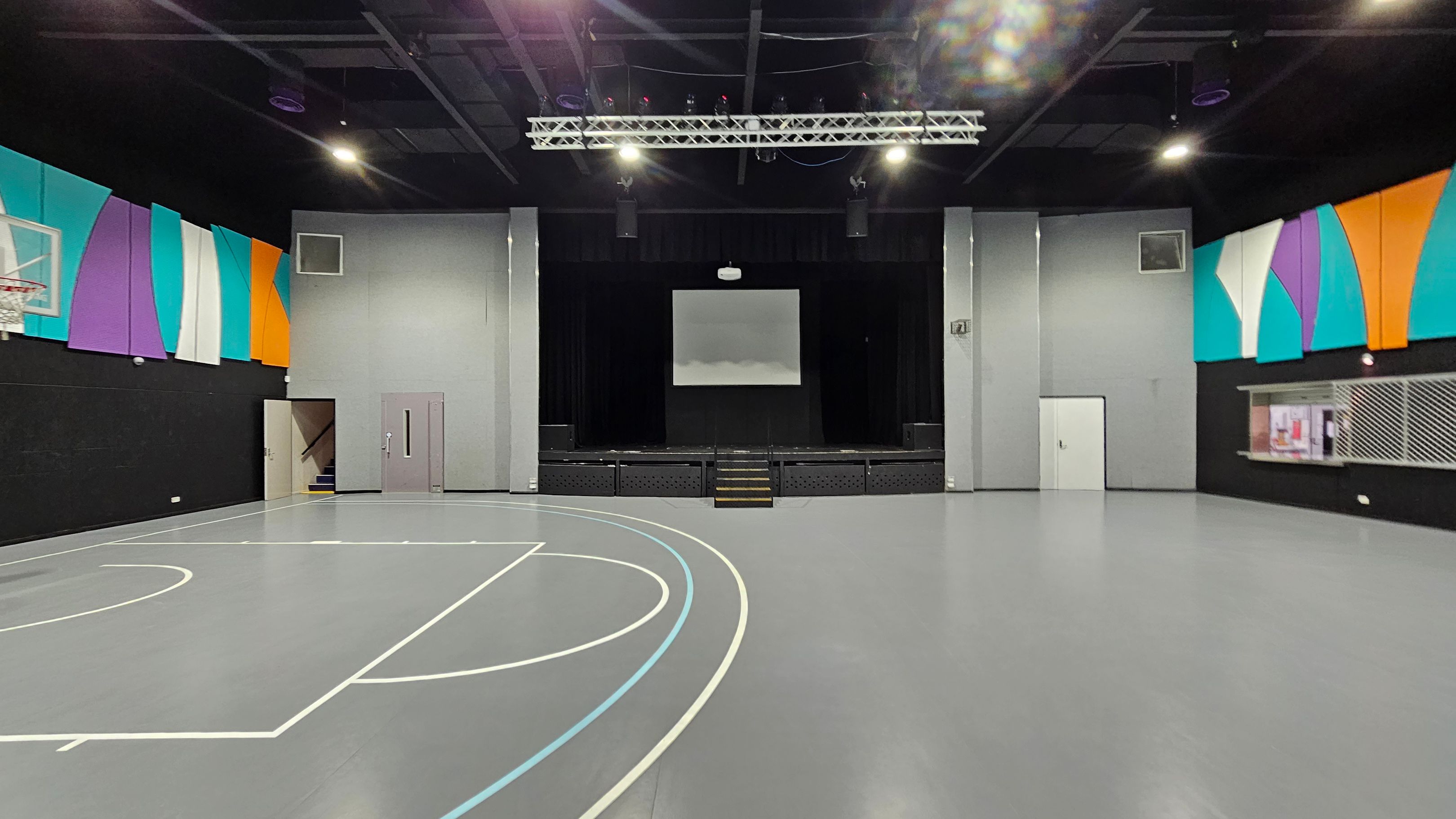 A spacious indoor gymnasium with a grey floor featuring basketball court lines. On the left, a basketball hoop is mounted on a wall adorned with colourful panels in teal, purple, white, and orange. In the centre, a stage with a screen above it is flanked by grey walls. On the right, a serving counter with a metal grille is set into the wall, which also has colourful panels. The ceiling is black with exposed beams and lighting fixtures.