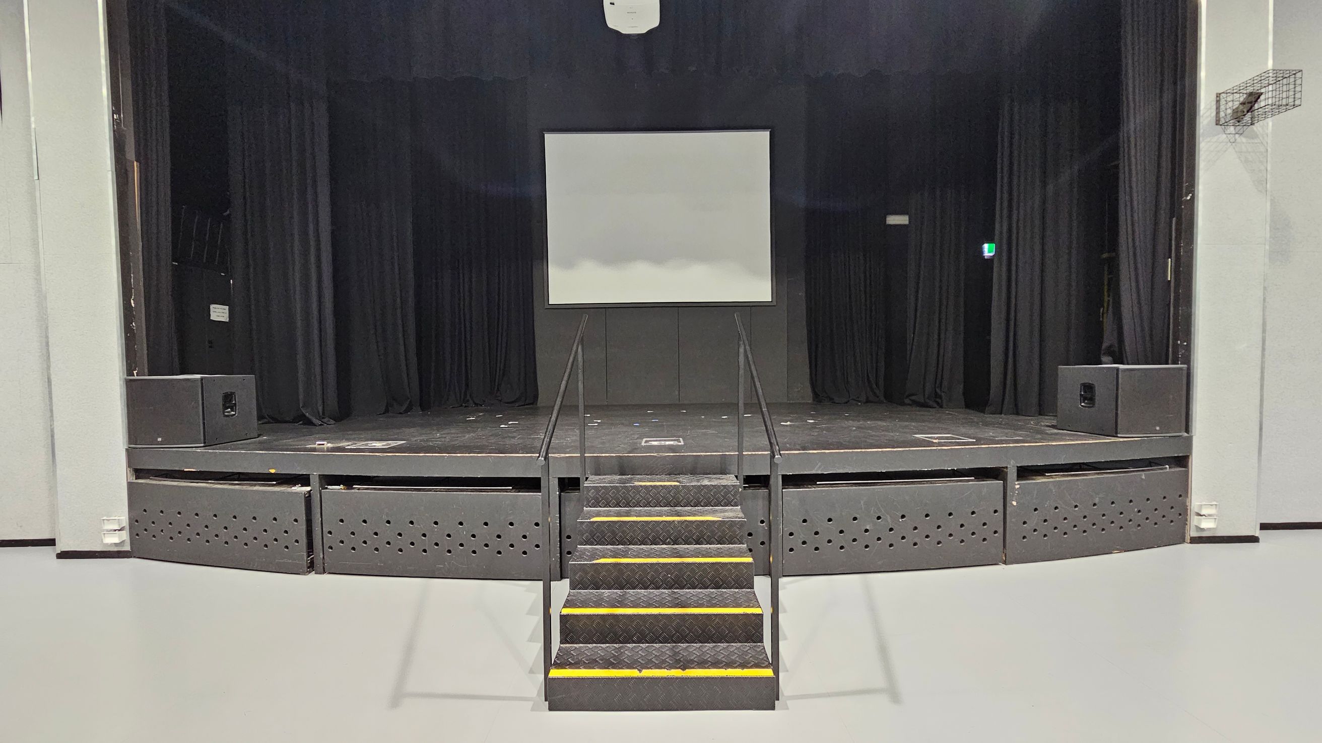 A stage with a central staircase leading up to it, featuring black railings. The stage has a large white screen in the middle, flanked by black curtains. Two large speakers are positioned on either side of the stage. The floor is light grey, and the ceiling is not visible. A small exit sign is illuminated on the right side.